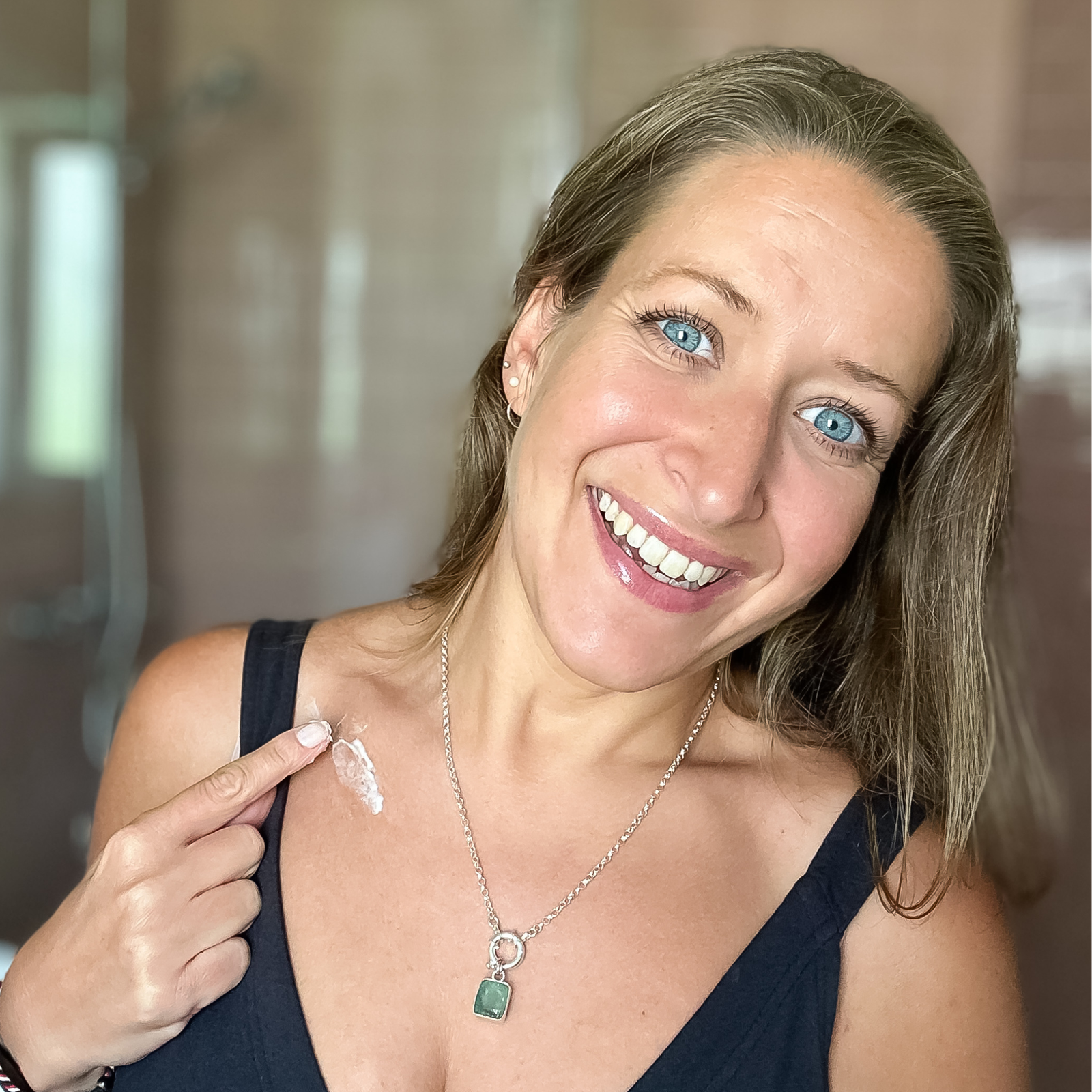 Woman smiling and applying CONCHUS natural skincare balm to her shoulder, in a bright bathroom setting.