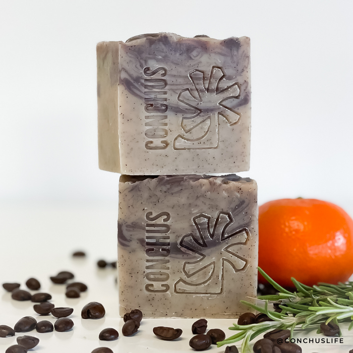 Two Olla exfoliating soap bars stacked with an orange, coffee beans and rosemary